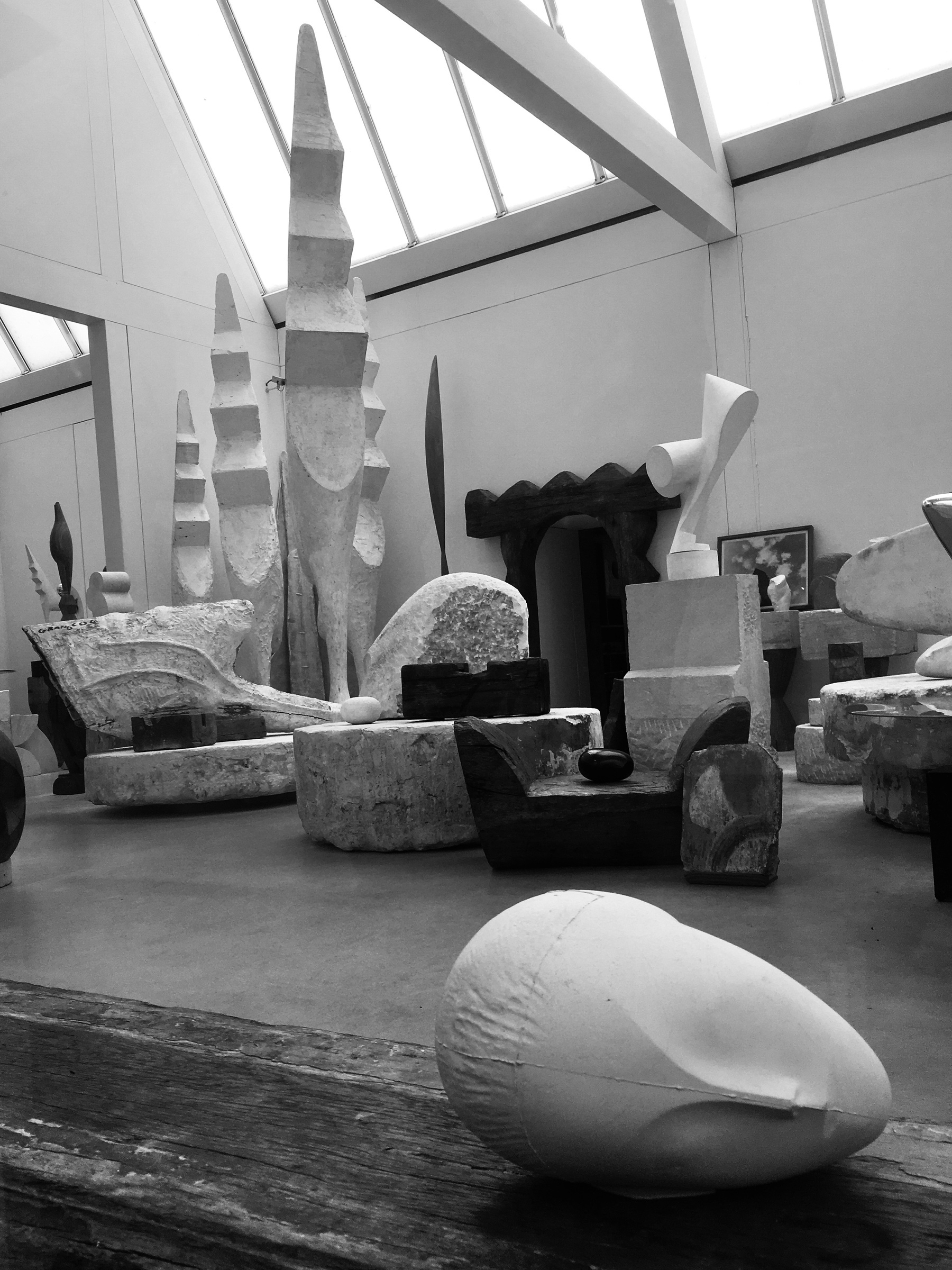 an black and white photo of the interior of the Atelier Brancusi in Paris. The sculptures Grand Coqs are in the corner of a room. Other sculptures surround them. In the foreground, a plaster sculpture of a head lies on a large block of wood. 